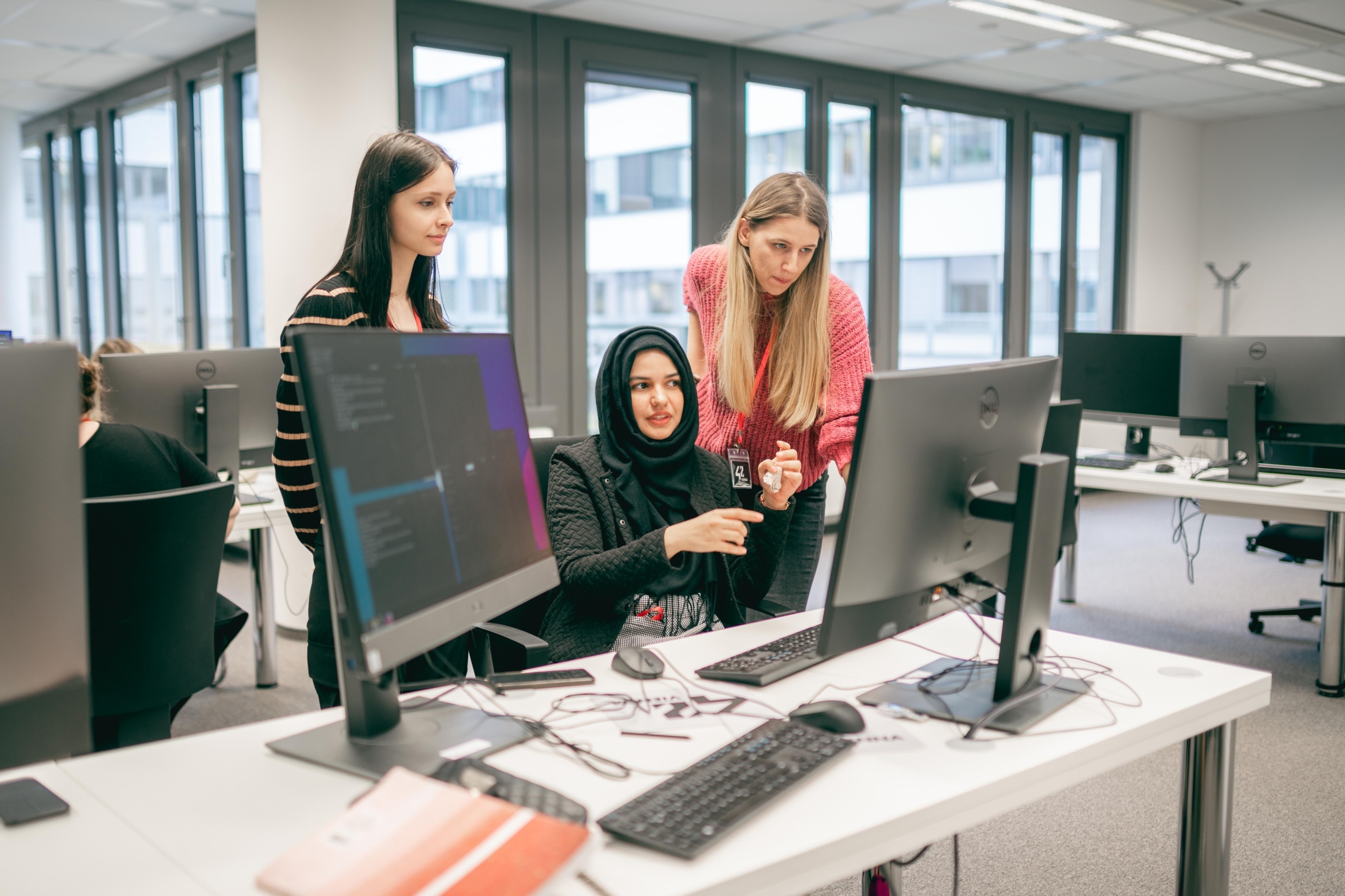 Eine Frau sitzt vor einem Computer und unterhält sich dabei mit zwei weiteren Frauen, die rechts und links neben ihr stehen und ebenfalls auf den gleichen Computerbildschirm sehen.